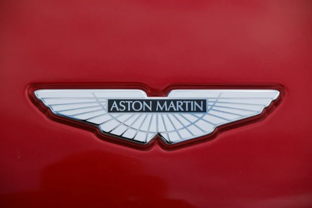 Aston Martin logotipas ant raudonos spalvos automobilio paviršiaus, simbolizuojantis prabangą ir greitį.