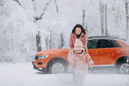 Mama su vaiku žaidžia sniege prie oranžinio automobilio, apsirengę rožine žiemine apranga žiemos miške.